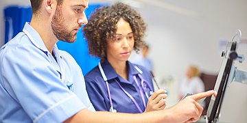 Foto: Ein Mann und eine Frau – beide in medizinischer Arbeitskleidung – stehen vor einem Computer, in den der Mann etwas eintippt.