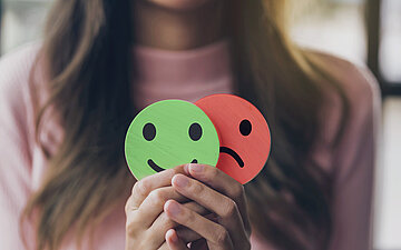 Foto: Eine Frau h&auml;lt in ihren H&auml;nden zwei Symbole &ndash; eines zeigt einen lachenden, das andere einen weinenden Smiley.