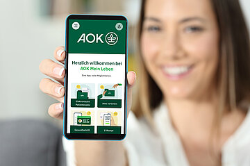 Foto zeigt etwas unscharf das l&auml;chelnde Gesicht einer Frau im Hintergrund, die mit einer Hand das Display ihres Smartphones in die Kamera h&auml;lt.