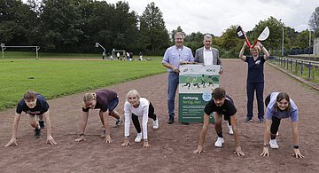 Startklar für den landesweiten Laufabzeichen-Wettbewerb Foto zeigt einen Schul-Sportplatz, auf der Aschebahn haben sich fünf Schülerinnen und Schüler in Startposition gebracht, dahinter stehen Schulleiter und AOK-Repräsentant mit dem Plakat zum Lauftag sowie rechts danaben die Vertreterin des SHLV mit einer Startklappe.