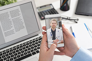 Ein Schreibtisch auf dem ein Laptop steht, auf dem Bildschirm ist ein Text sichtbar. Im Hintergrund B&uuml;routensilien, wie zum Beispiel ein Taschenrechner. Im Vordergrund halten zwei H&auml;nde ein Smartphone, auf dem ein Arzt zu sehen ist.