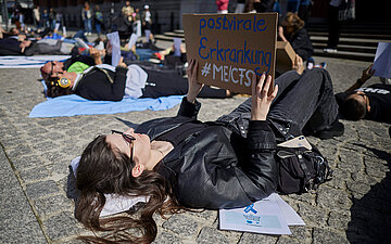 Foto einer jungen Frau, die im Liegen demonstiert mit einem Plakat in den H&auml;nden, auf dem "Postvirale Erkrankung ME/CFS steht"