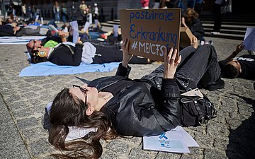 Fatigue-Syndrom Foto einer jungen Frau, die im Liegen demonstiert mit einem Plakat in den Händen, auf dem "Postvirale Erkrankung ME/CFS steht"