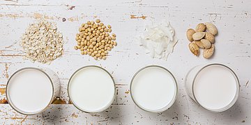 Milchersatzprodukte Foto: Kleine Schälchen mit Milchähnlichen Flüssigkeiten, davor liegen verschiedene Getrieide, Reis oder Bohnen.