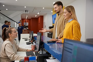 forsa-Umfrage zur Primärversorgung in Westfalen-Lippe In einer Praxis steht ein junges Paar am Empfangstresen und ist im Gespräch mit einer medizinischen Angestellten, die auf der anderen Seite des Tresens sitzt.