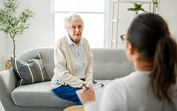 Eine ältere Frau sitzt auf auf der Sofakante eines Wohnzimmers, ihr gegenüber mit dem Rücken zum Betrachter sitzt eine Frau mit Klemmbrett und Stift in der Hand und stellt Fragen.