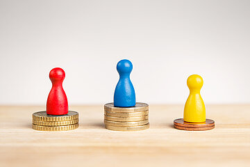 Foto: Drei Spielfiguren in den Farben Rot, Blau und Gelb stehen auf M&uuml;nzstapeln, die unterschiedlich hoch sind.