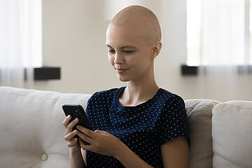 Eine junge Frau mit Glatze sitzt auf dem Sofa und schaut auf ihr Handy.