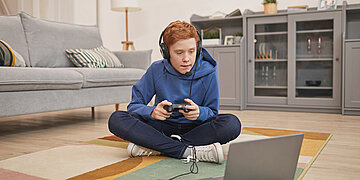Foto: Ein Junge sitzt mit einem Controller und Kopfh&ouml;rern im Schneidersitz vor einem Laptop.