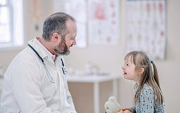 Versorgung Behinderte Doktor sitzt neben einem Mädchen im Behandlungszimmer. Das Kind hält einen Teddybär.