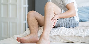 Foto: Ein Mann sitzt auf dem Bett und fasst sich an die linke Wade.