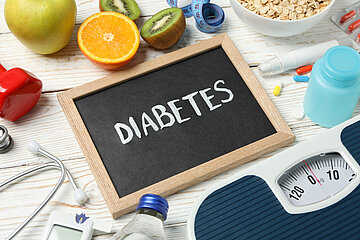 Auf einem Tisch liegt eine kleine Schiefertafel auf dem das Wort 'Diabetes' steht. Kreisf&ouml;rmig darum herum angeordnet sind Obst, Ma&szlig;band, Medikamente, eine K&ouml;rperwaage, Teil einer Getr&auml;nkeflasche, ein Stethoskop, ein Blutzuckermessger&auml;t und St&uuml;ck einer roten Hantel.