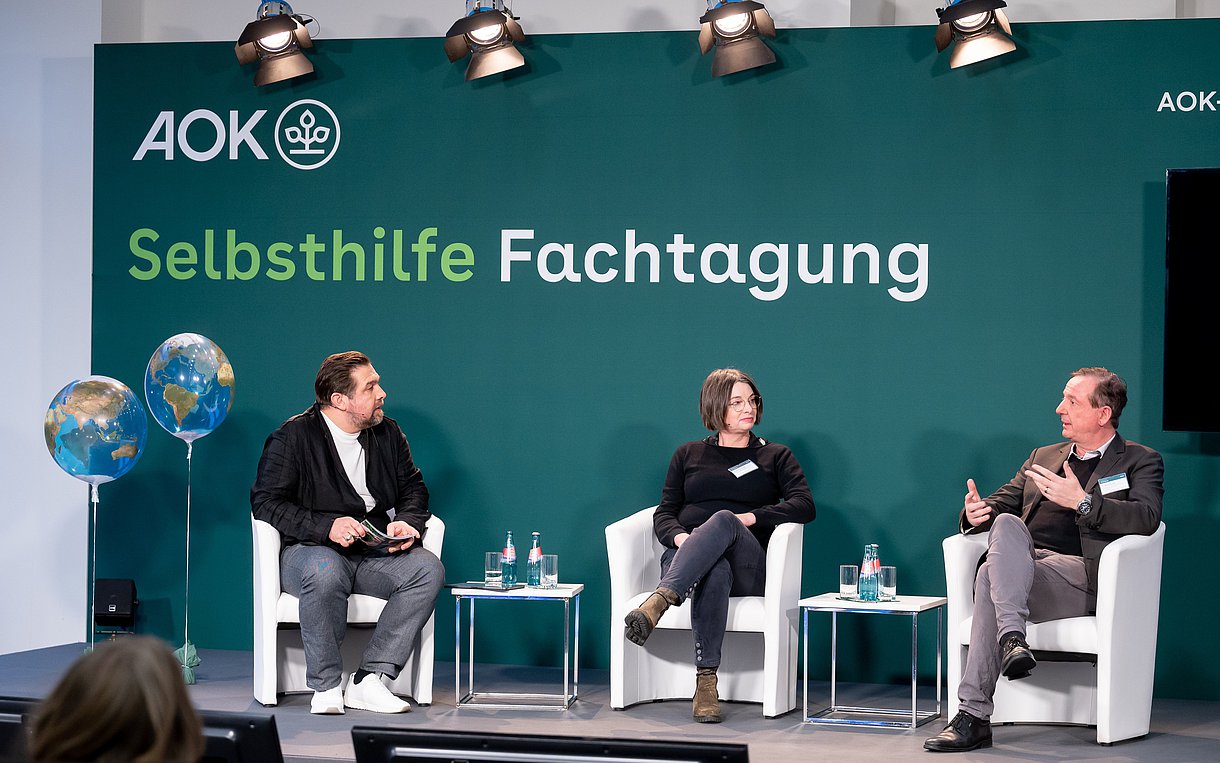 Moderator Michael Bernatek und die Referentinnen und Referenten Dr. Annette Scheder und Dr. Martin Danner sitzen in Sesseln auf einer Bühne, auf zwei Bildschirmen rechts von ihnen sind Maike Voss sowie Prof. Dr. Claudia Traidl- Hoffmann zu sehen.