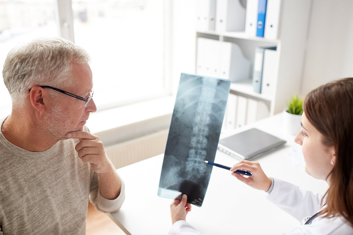 Foto zeigt ein Arzt-Patientengespräch, bei der eine Ärztin einem Patienten ein Röntgenbild mit einer Wirbelsäule zeigt und erklärt.