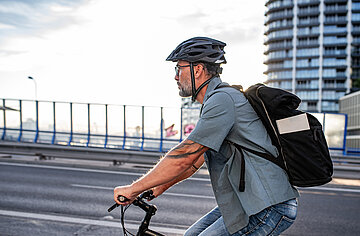 Ein Fahrradfahrer f&auml;hrt mit seinem Rad auf einer ansteigenden Br&uuml;ckenstra&szlig;e; vom Rad ist nur der Lenker sichtbar ist. Der Mann tr&auml;gt einen Fahrradhelm und hat einen Rucksack auf dem R&uuml;cken. Im Hintergrund Br&uuml;ckengel&auml;nder und die verglaste Frontfl&auml;che eines Hochhauses.