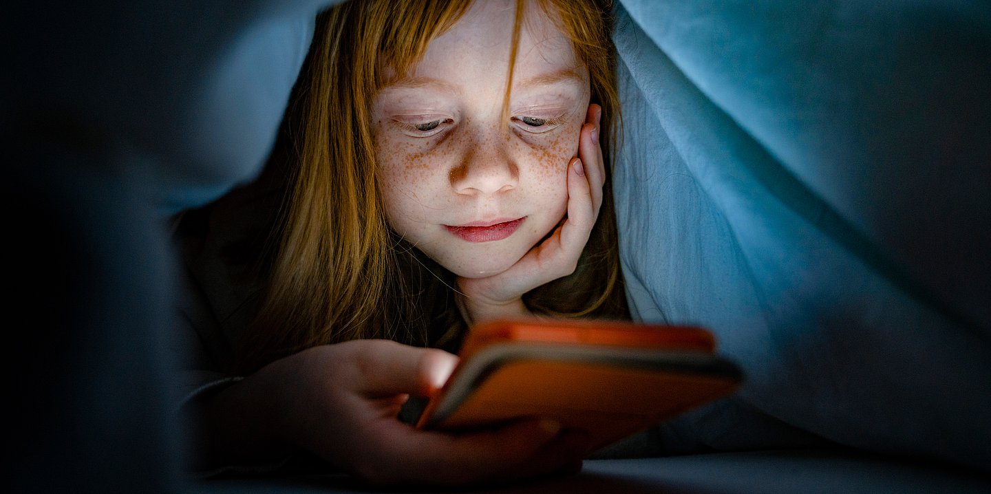 Foto: Ein Kind mit Decke über dem Kopf schaut auf ein Smarthphone, das es in der Hand hält.