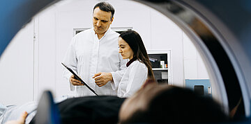 Zwei medizinische Fachkr&auml;fte besprechen Dokumente vor einem MRT-Ger&auml;t, in dem eine Person untersucht wird. 
