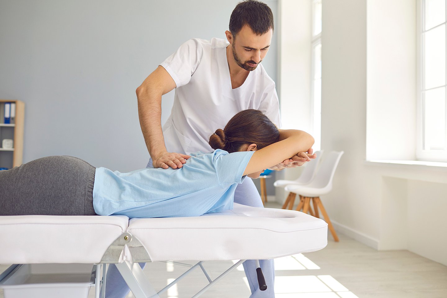 Foto zeigt einen Physiotherapeuten, der eine Frau, die auf einer Behandlungsliege platziert ist, physiotherapeutisch behandelt.