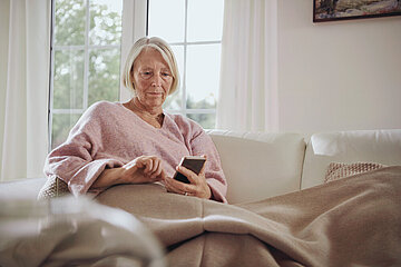 Symbolbild einer &auml;lteren Frau, die auf einem Sofa sitzt und ihr Smartphone antippt.