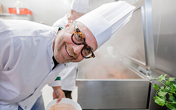 Koch mit wei&szlig;er M&uuml;tze arbeitet in einer Gro&szlig;k&uuml;che und l&auml;chelt in die Kamera.