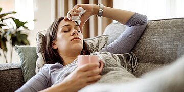 Das Bild zeigt eine Junge Frau, die offenbar krank und ersch&ouml;pft auf einer grau gemusterten Couch liegt. In der rechten Hand h&auml;lt sie eine pinkfarbene Teetasse. Ihre linke Hand h&auml;lt ein Taschentuch und liegt an der Stirn. Im Hintergrund ist ein Fenster mit Zimmerpflanzen zu sehen.