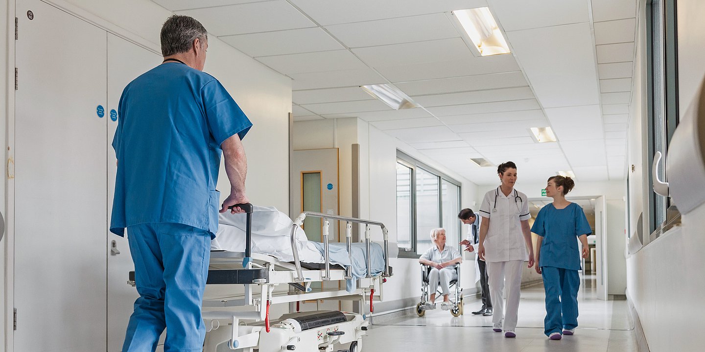 Foto: Ein Pfleger in blauer Tracht schiebt ein Krankenhausbett über einen Klinikflur.