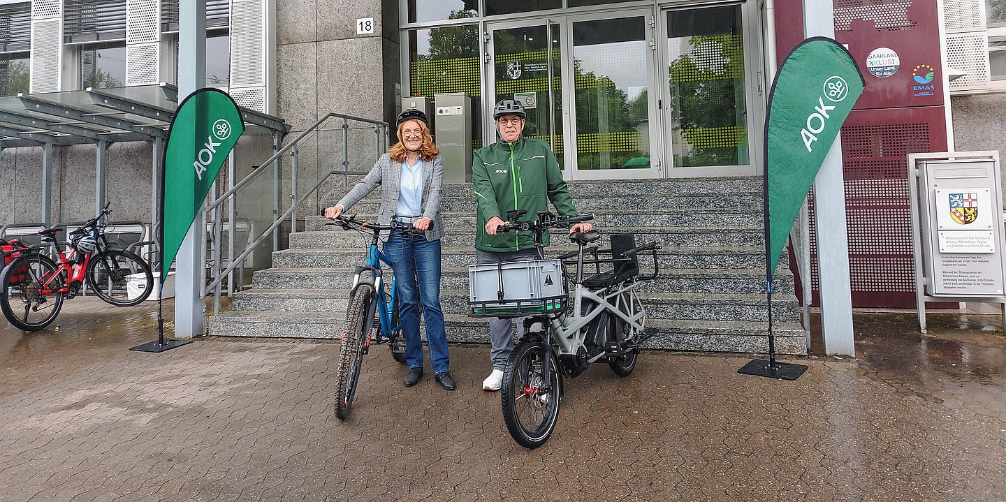 Foto zeigt eine Frau (links) mit Helm und Fahrrad und einem Mann (rechts) mit Helm und Fahrrad vor einem Verwaltungsgebäude