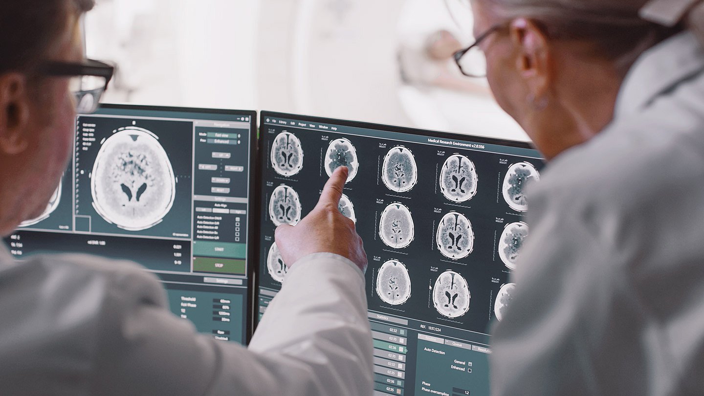 Foto: Zwei Ärzte beugen sich über Hirnscans am Computerbildschirm, sie scheinen Auffälligkeiten zu besprechen.