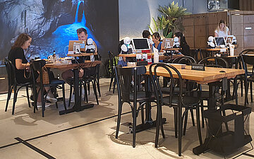 Foto von G&auml;sten des Robotercaf&eacute;s. Auf den Tischen befinden sich Computer und kleine Miniaturroboter.