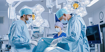 In einem Operationssaal stehen zwei &Auml;rzte und eine Krankenschwester in OP-Kitteln und operieren einen Patienten.