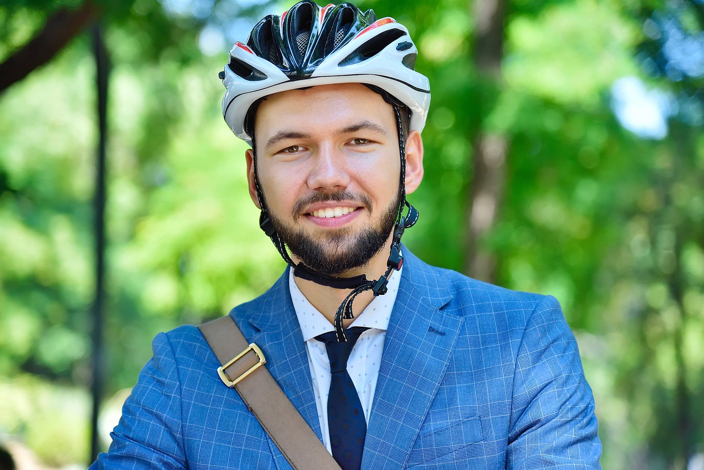 Foto zeigt einen jungen Mann in einem blauen Anzug, darunter Schlips und Hemd, der einen Fahrradhelm trägt und fahrbereit aussieht. Im Hintergrund ist Wald zu erkennen..