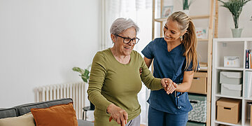 Eine junge weibliche Pflegekraft hilft einer &auml;lteren Dame beim Laufen am Stock im h&auml;uslichen Umfeld.