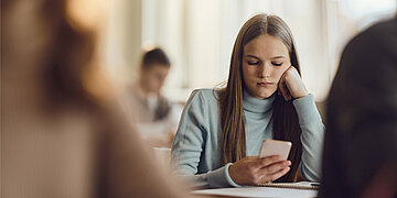 Eine Sch&uuml;lerin schaut w&auml;hrend des Unterrichts gelangweilt auf ein Smartphone.