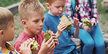 Mehrere Kinder sitzen drau&szlig;en zusammen und essen Sandwiches.