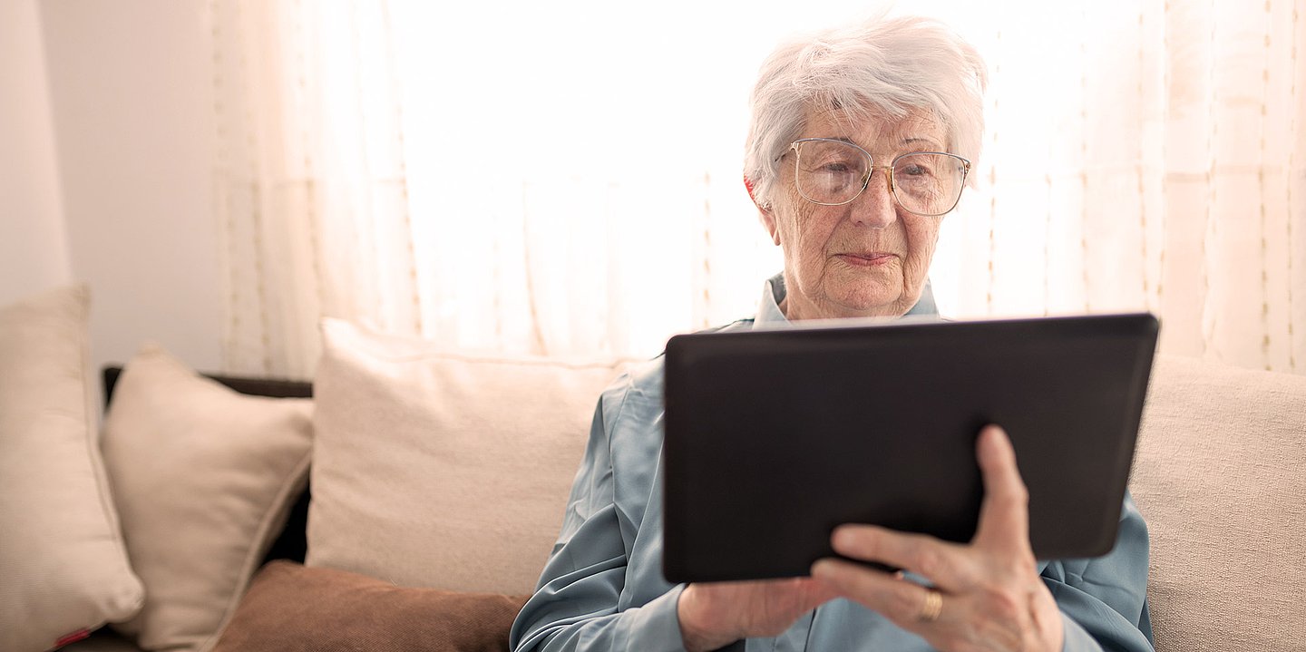 Foto: Eine ältere Frau sitzt auf dem Sofa und hält ein Tablet in der Hand.