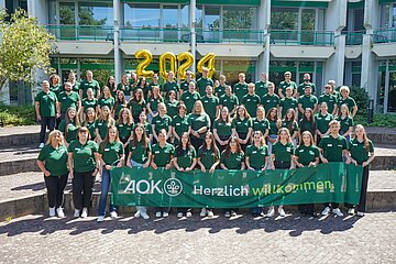Das Gruppenbild zeigt den Ausbildungsjahrgang 2024. Die Azubis in vier aufsteigenden Reihen tragen alle das gr&uuml;ne AOK-Poloshirt. Die vorderste Reihe h&auml;lt ein Banner mit dem AOK-B&auml;umchen und der Aufschrift "Herzlich Willkommen". Oberhalb der Gruppe h&auml;ngen goldene Luftballons, die die Jahreszahl 2024 bilden.