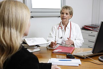 Das Bild zeigt, wie eine &Auml;rztin an ihrem Schreibtisch mit einer Patientin spricht.