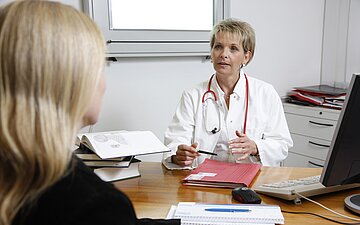Das Bild zeigt, wie eine &Auml;rztin an ihrem Schreibtisch mit einer Patientin spricht.