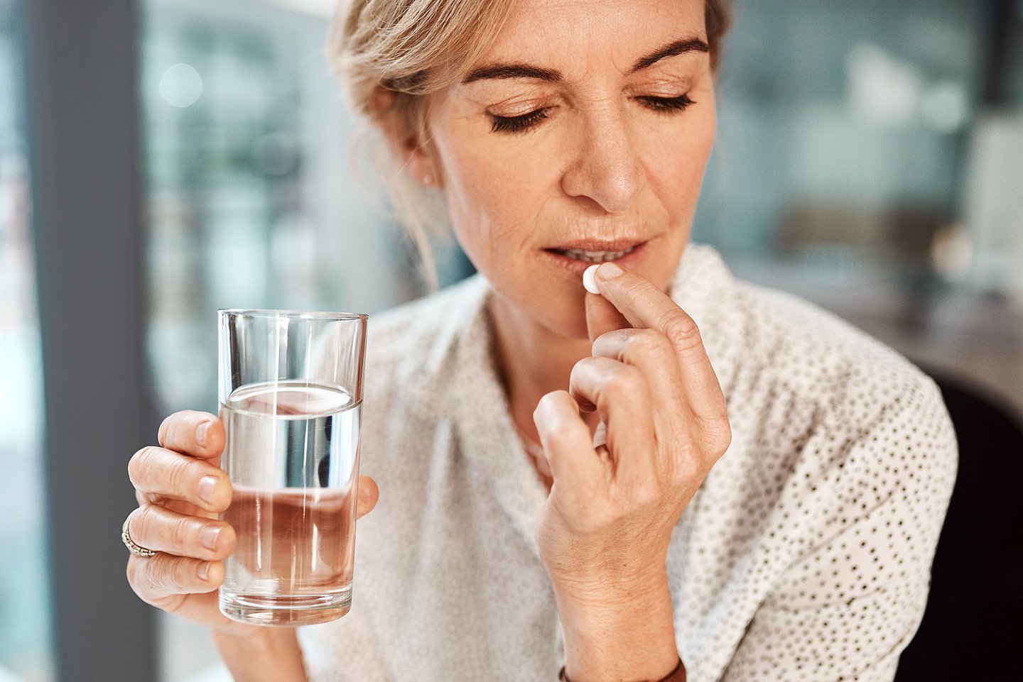 Foto einer Frau, die eine Tablette einnimmt, dazu ein Glas Wasser in der Hand.