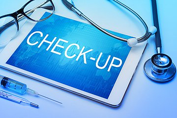 Gesundheits-Check-up in Westfalen-Lippe Ein Laptop mit dem Schriftzug 'CHECK-UP' auf dem Display liegt auf einem Tisch, umrahmt von Brille, Stethoskop, Spritze und Fieberthermometer.