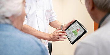 Ein Arzt zeigt zwei &auml;lteren Personen auf einem Tablet eine Ern&auml;hrungspyramide w&auml;hrend eines Beratungsgespr&auml;chs in der Praxis.
