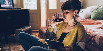 Alkoholkonsum Foto: Eine Frau schaut auf ihr Tablet und trinkt dabei Rotwein.