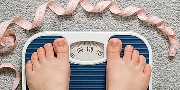 Übergewicht (bei Kindern) Das Foto zeigt die Zehen von zwei Füßen auf einer Personenwaage. Die analoge Anzeige zeigt 105 Kilogramm. Über der Waage liegt ein weißes Maßband mit roter Skala in Spiralen ge- beziehungsweise verdreht.