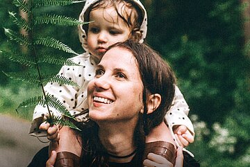 Eine Mutter tr&auml;gt ihr Kind auf ihren Schultern durch einen Wind. Links ist ein Farnzweig zu sehen.