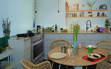 Das Bild zeigt die Küche der Musterwohnung Demenz. Sie ist wie eine richtige Küche mit verschiedenen Alltagsgegenständen ausgestattet. Zu sehen sind ein gedeckter Tisch mit vier Plätzen sowie im Hintergrund eine Küchenzeile mit Herd und Spülbecken.