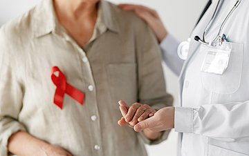 HIV Foto einer älteren Dame mit HIV-Schleife, die eine Medizinerin an der Hand hält. Die Gesichter sind nicht zu sehen.