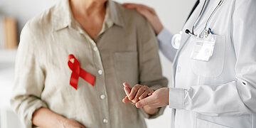 Foto einer &auml;lteren Dame mit HIV-Schleife, die eine Medizinerin an der Hand h&auml;lt. Die Gesichter sind nicht zu sehen.