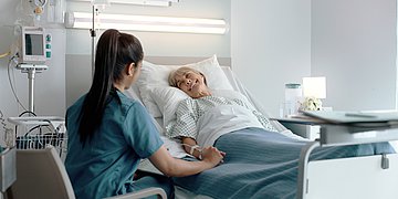 Klinikzufriedenheit In einem Krankenhausbett liegt eine ältere Dame lächelnd und hält die Hand einer vor dem Bett sitzenden Pflegerin.
