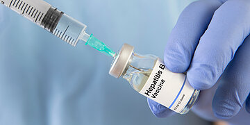 Foto: Eine behandschuhte Hand h&auml;lt ein Fl&auml;schchen mit Hepatitis-B-Impfstoff, w&auml;hrend eine Spritze in den Verschluss eingef&uuml;hrt wird.