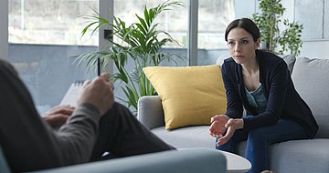 PINAH für Patientinnen und Patienten aller Krankenkassen Das Foto zeigt eine Frau, auf einer Coach sitzend, im Gespräch mit einer ihr gegenübersitzenden männlichen Person, die sich dabei Notizen macht.